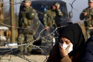 Pietà - Νεαρή γυναίκα, ντυμένη στα μαύρα, κρατά μια μάσκα στο πρόσωπό της, έχοντας καθίσει αποκαμωμένη δίπλα στο αγκαθωτό σύρμα των συνόρων υπό το βλέμμα των στρατιωτών της ΠΓΔΜ, που αναμένουν εντολές στην άλλη πλευρά (Ειδομένη, 19 Νοεμβρίου 2015)