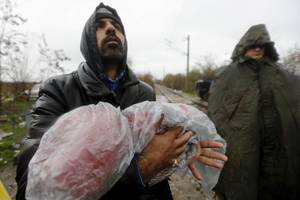 Η Αγία Τριάδα – Ιρανός κρατά το παιδί του, αναζητώντας να πιαστεί από κάπου μέσα στη βροχή υπό το άγρυπνο βλέμμα των φρουρών (Ειδομένη, 27 Νοεμβρίου 2015)